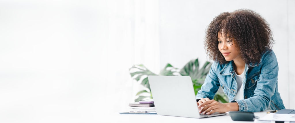 young woman sitting at a table at her laptop with books at her side and a plant in the background