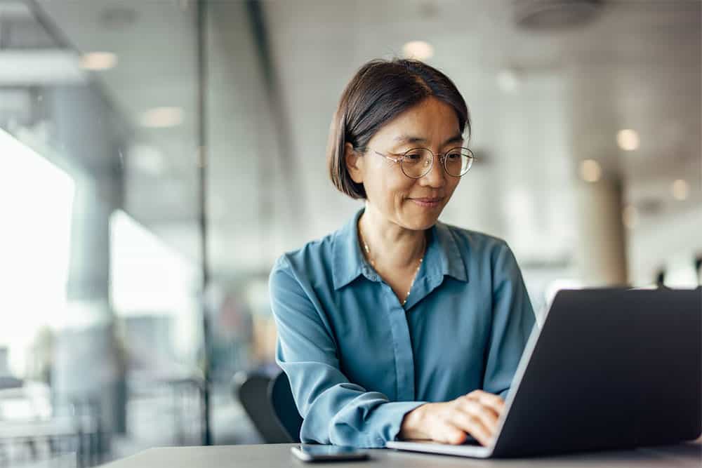 woman at her laptop sitting at a table in a room with floor to ceiling windows